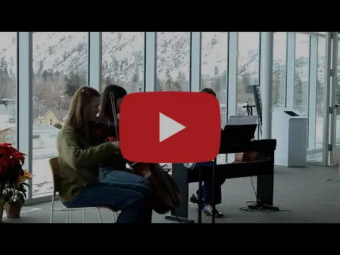 Two musicians play instruments in a room with large windows overlooking a snowy landscape.