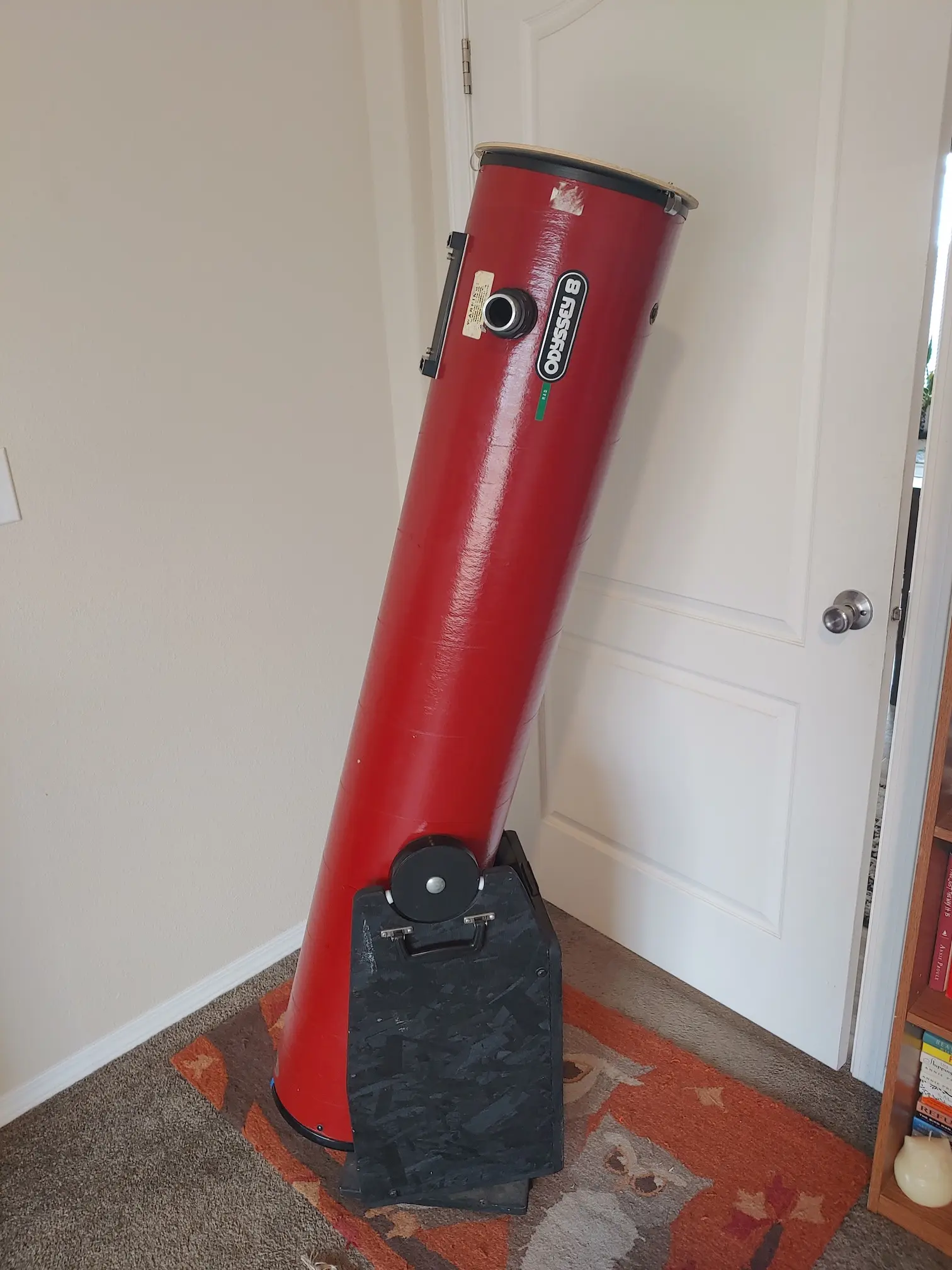 A red reflecting telescope stands on a patterned rug in a room.