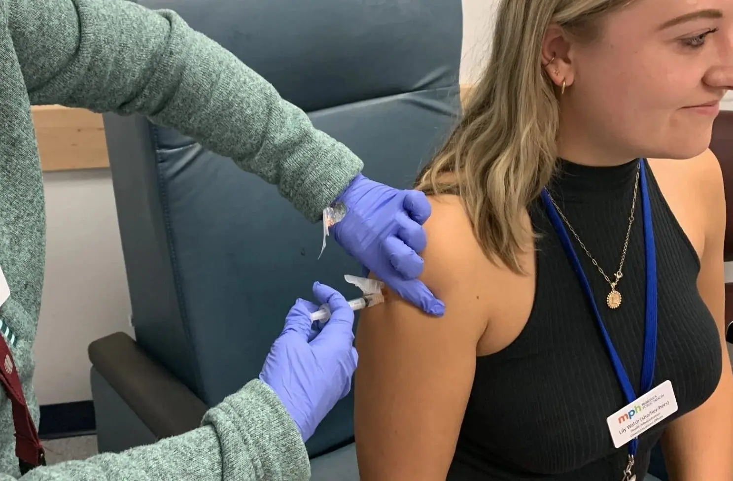A person receives an injection in their arm from a medical professional wearing blue gloves.