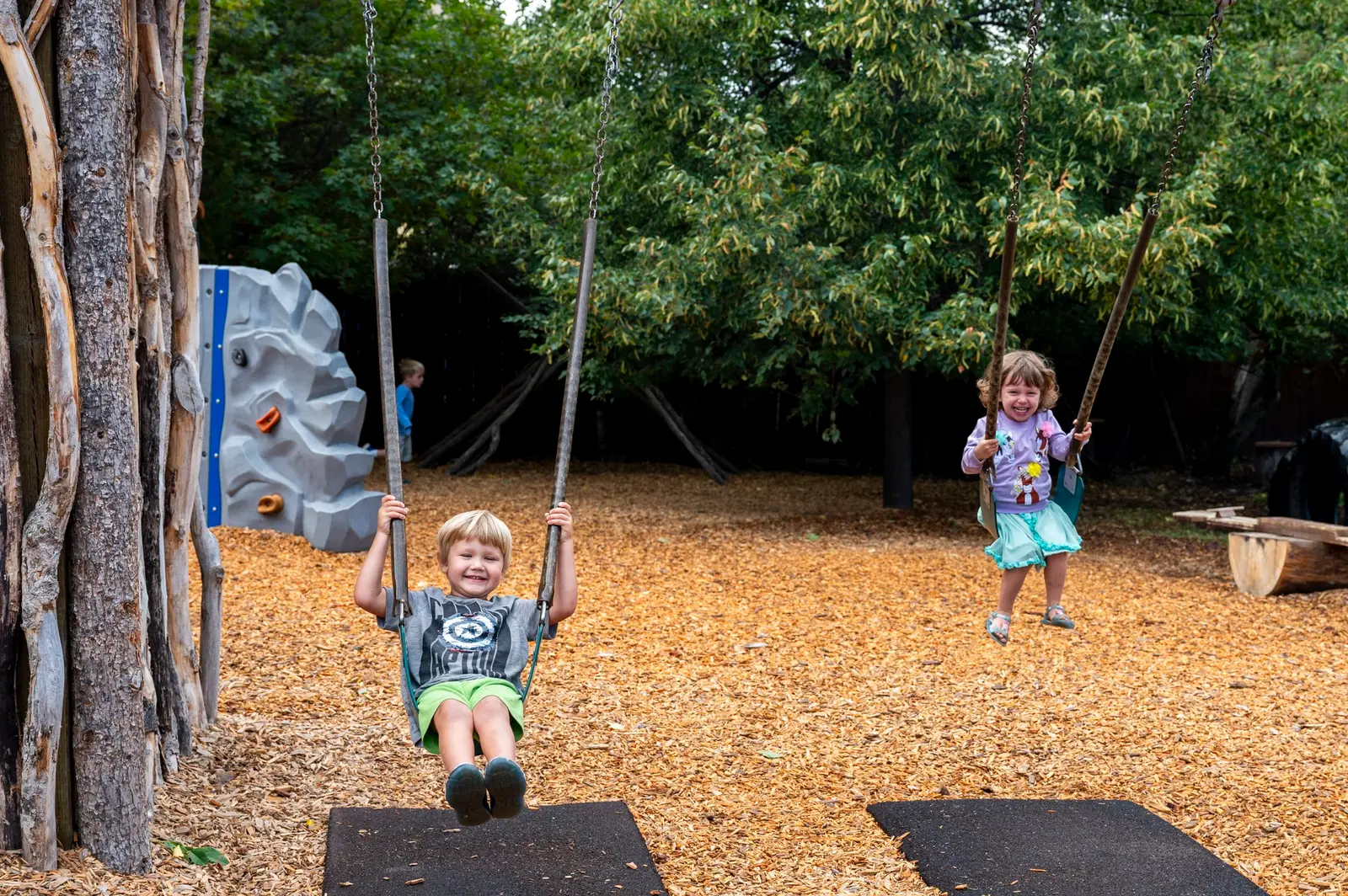 Two children swing on a playground while another child stands in the background.