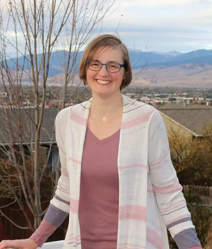 A smiling woman stands outdoors with a town and mountain landscape in the background.