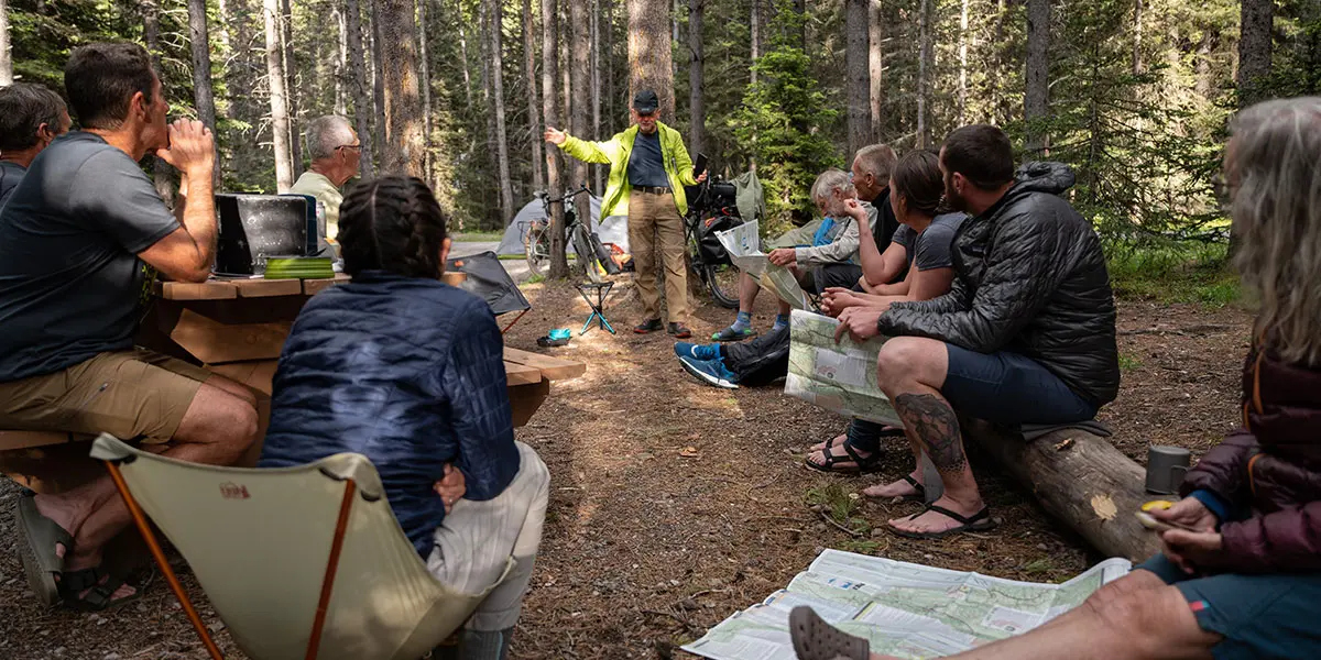 A group of adults sits in a forest clearing listening to an instructor talk about a map.