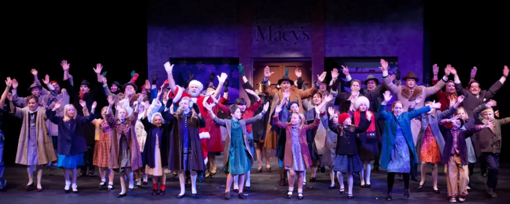 A large ensemble cast performs on stage in front of a backdrop depicting Macy's.