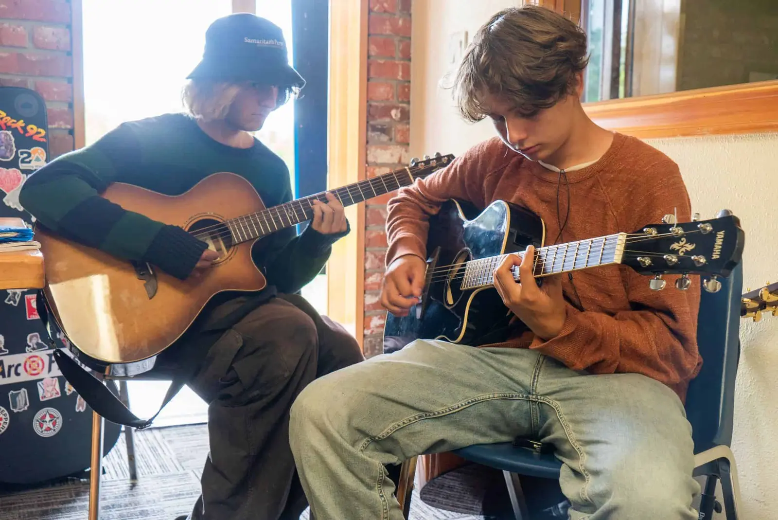 Two teenagers are playing acoustic guitars while sitting indoors.