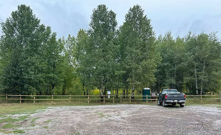 A pickup truck is parked in a gravel lot in front of a wooden fence and a grove of trees.