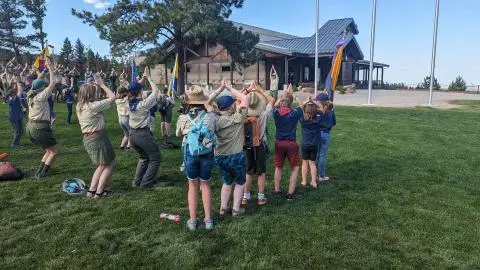A group of young people in scout uniforms stand on a lawn and hold their arms in the air.