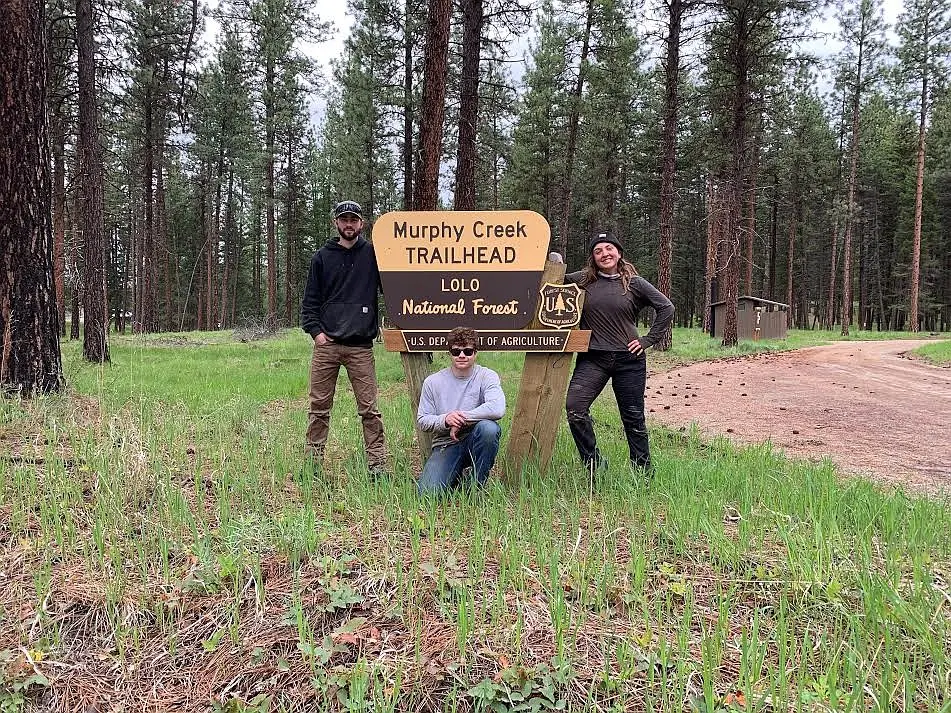 Three young adults stand and crouch in front of a forest trailhead sign.