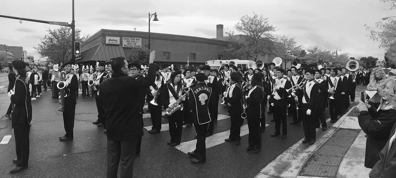 A marching band stands in formation on a city street while an instructor leads them.