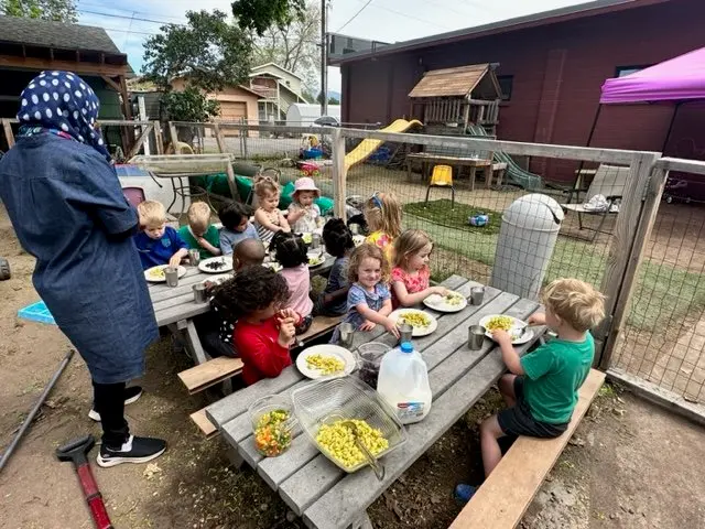 Children sitting at a long outdoor picnic table eating food, with a woman supervising, in a backyard with play equipment and a fenced area.