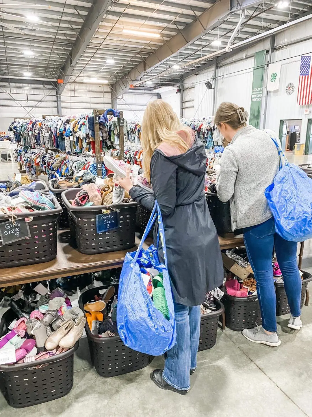 Two women shopping for shoes at a thrift store, with racks of clothing and baskets of shoes.