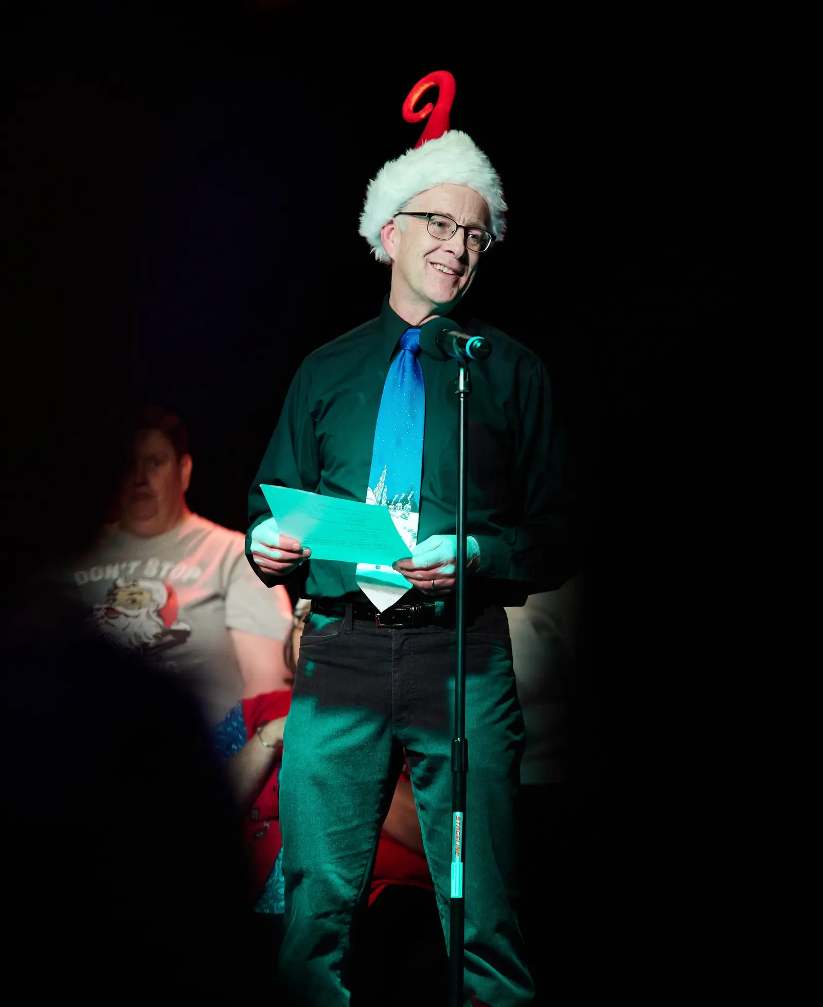 Man wearing a Santa hat with a red number 2, glasses, black shirt, blue tie, and holding papers, speaking into a microphone on stage, with a woman in a Don’t Stop T-shirt in the background.