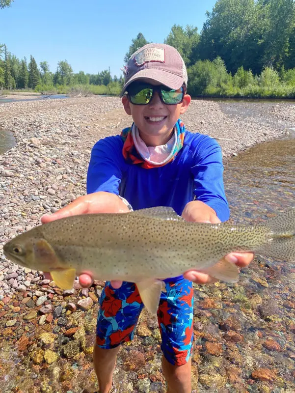 A child wearing sunglasses and a cap holds a caught fish in a stream.