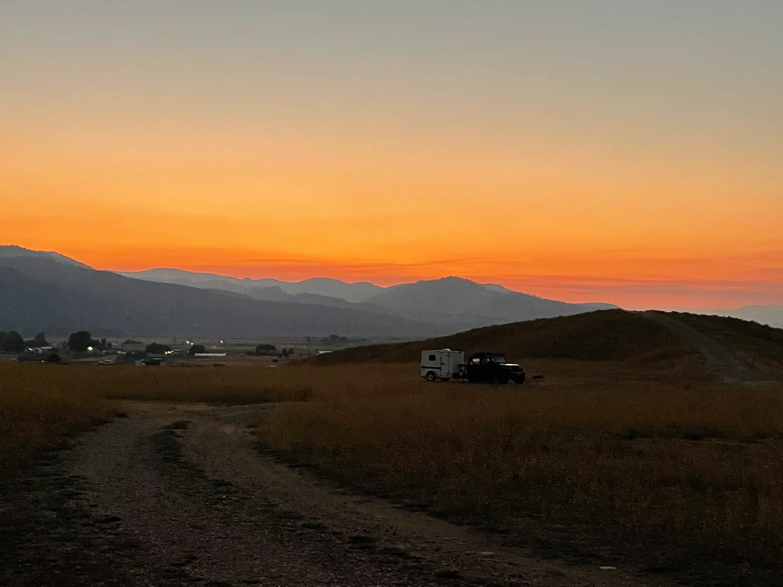 A vehicle with a small trailer sits in a field at sunset beneath a dramatic orange sky and mountains.