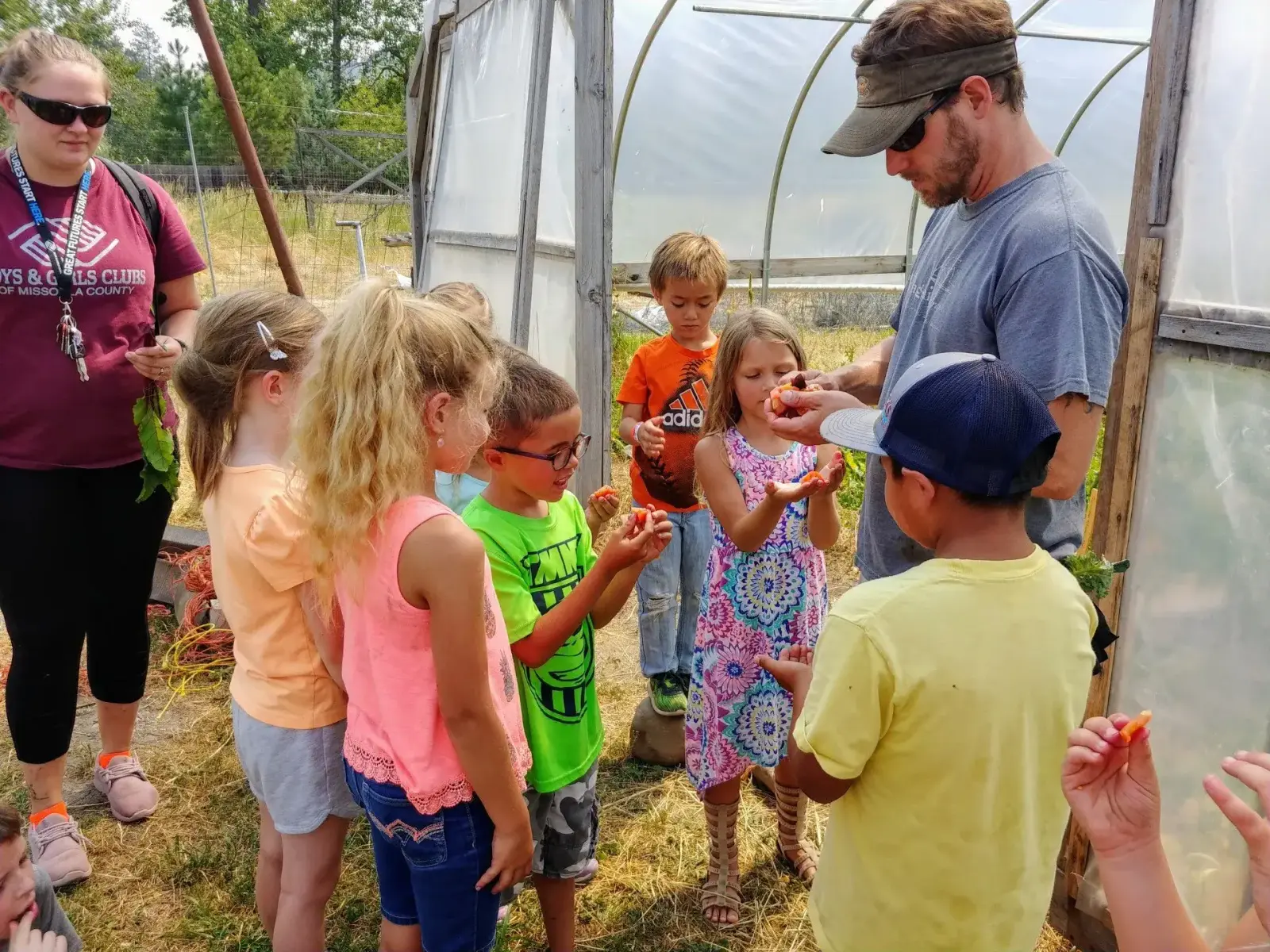 An adult guides a group of children as they examine produce outside a greenhouse.