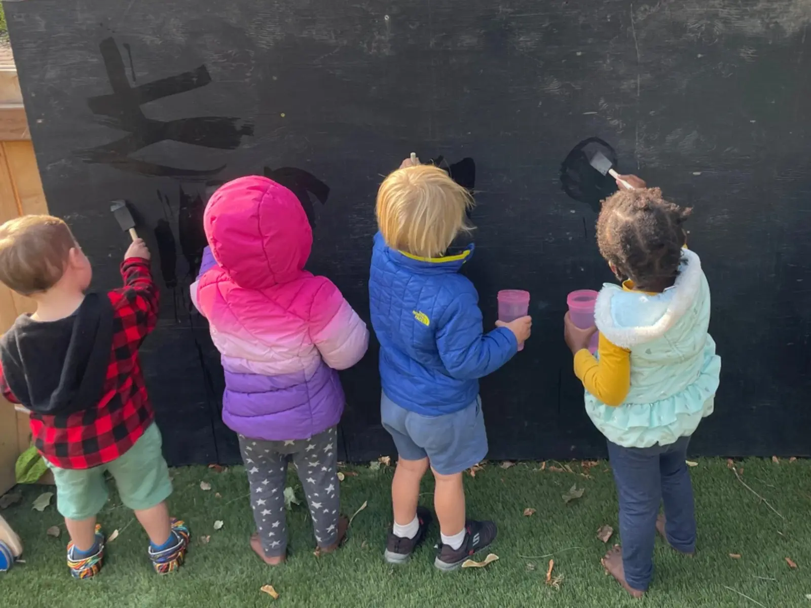Four children wearing colorful jackets are painting on a black wall with brushes. They are holding pink cups and are standing on green grass with some fallen leaves.