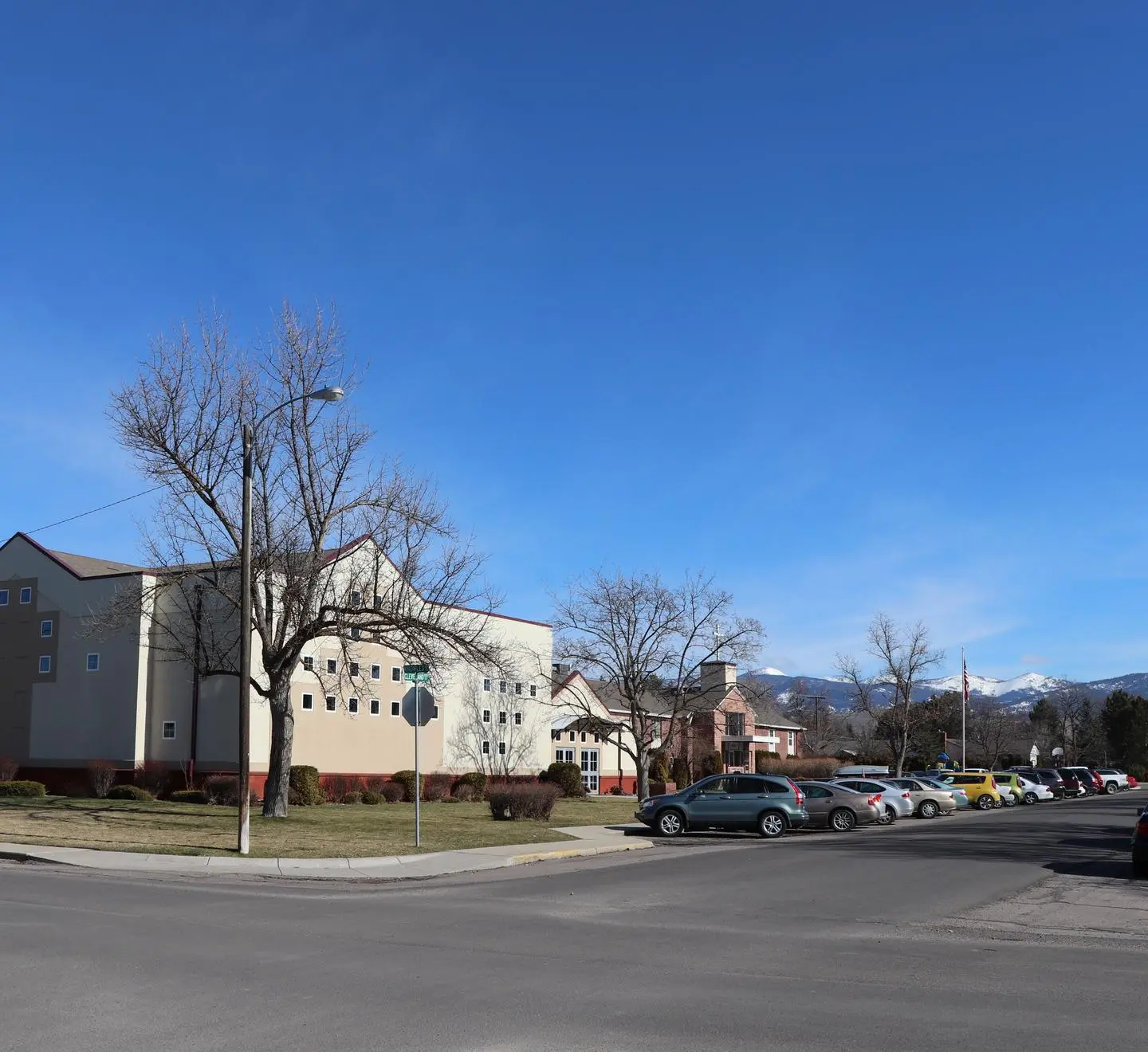 A street view shows buildings and a row of cars parked under a bright blue sky with mountains in the background.