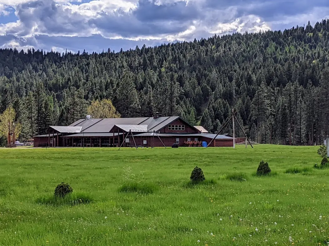 A large wooden building sits at the edge of a meadow with a dense forest in the background.