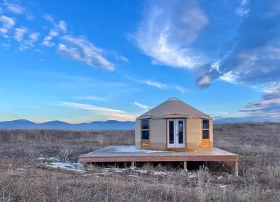 Photo from primrosestation.com A round yurt sits on a wooden deck in a grassy field with mountains in the background.