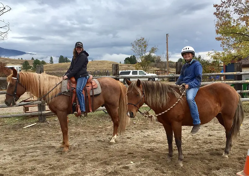 A woman and a young person are riding horses in a sandy paddock.