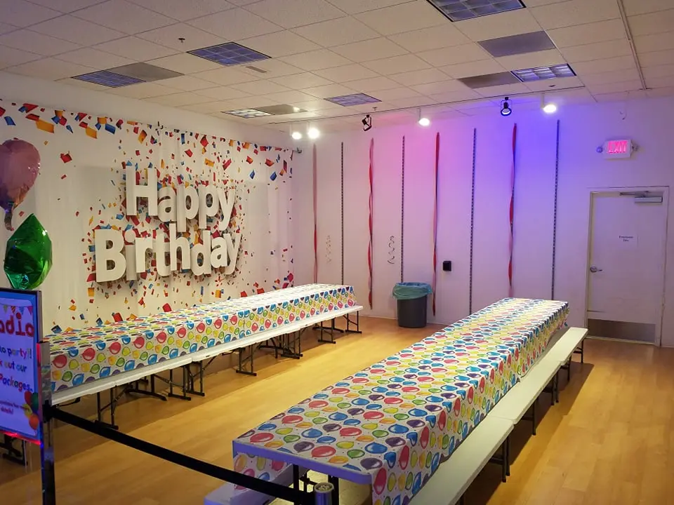 An indoor party room is decorated with a Happy Birthday sign and tables with patterned tablecloths.