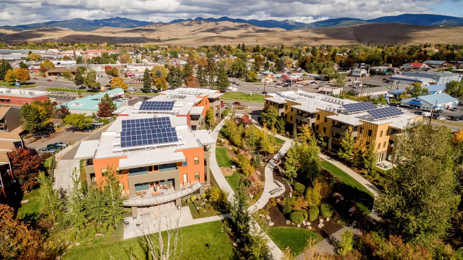 An aerial view shows modern multi-story buildings with rooftop solar panels surrounded by landscaping and a small town.