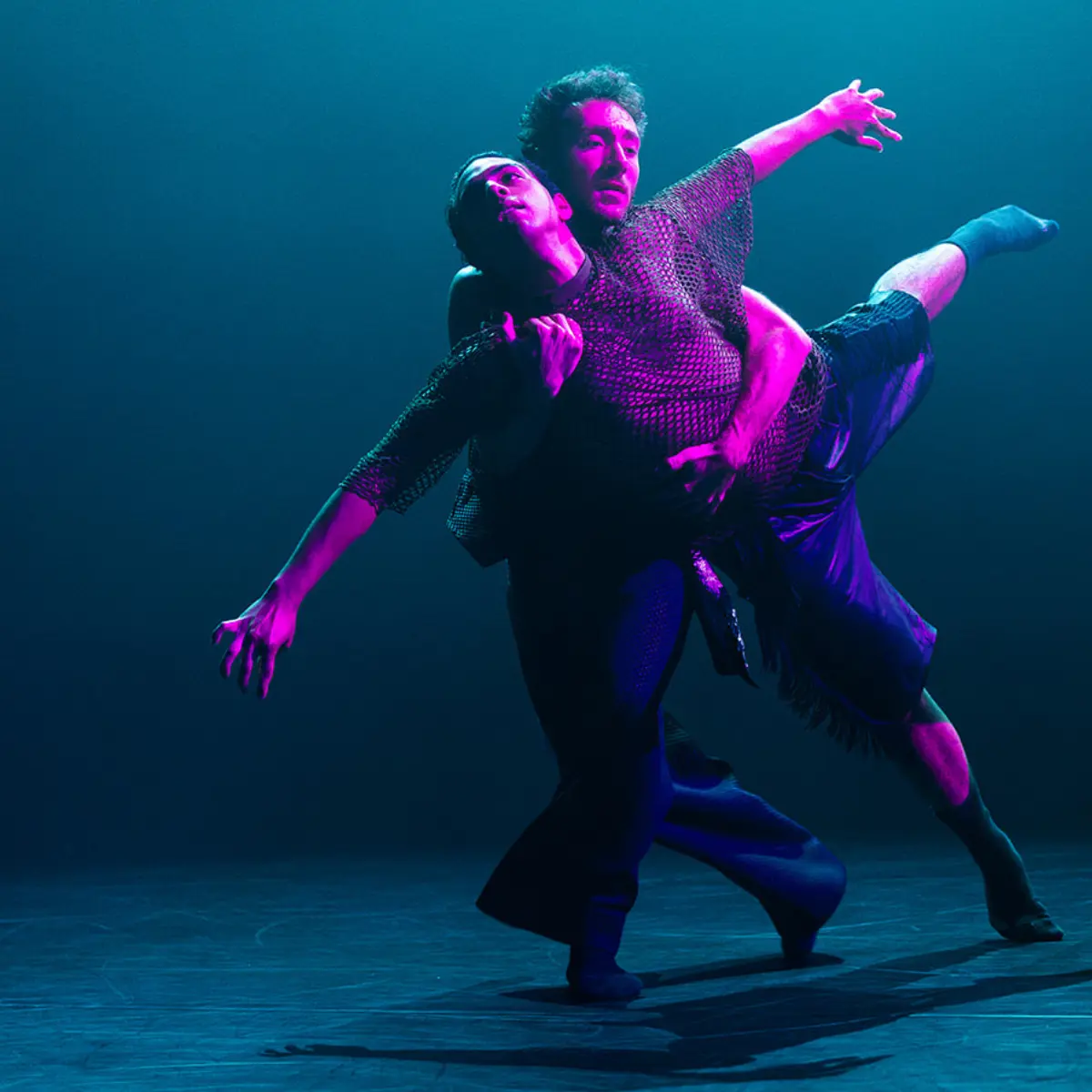 Two performers in mesh shirts are dancing together under blue and pink stage lights.
