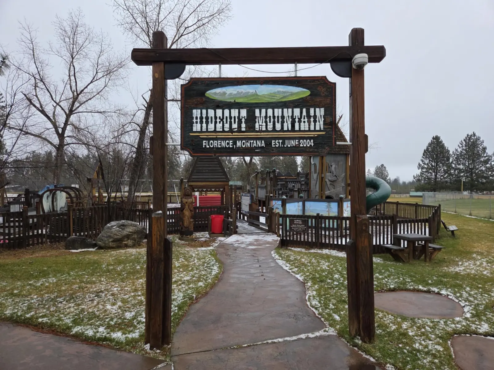 A wooden sign for a playground stands in an outdoor area with light snow.