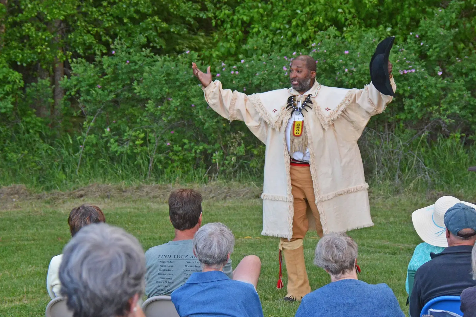A man in traditional attire stands with his arms open while speaking to a seated audience outdoors.
