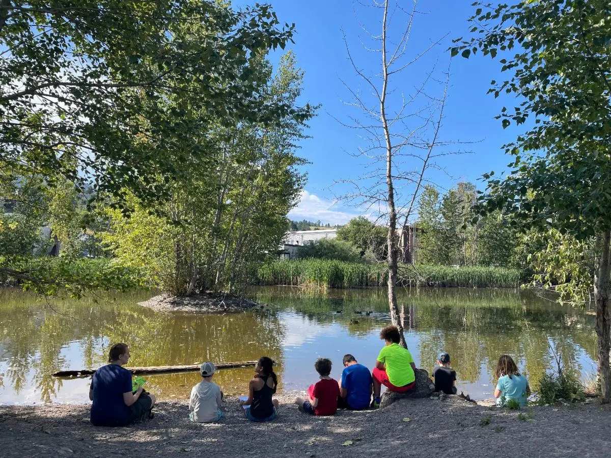 A group of children and an instructor sit by a pond outdoors.