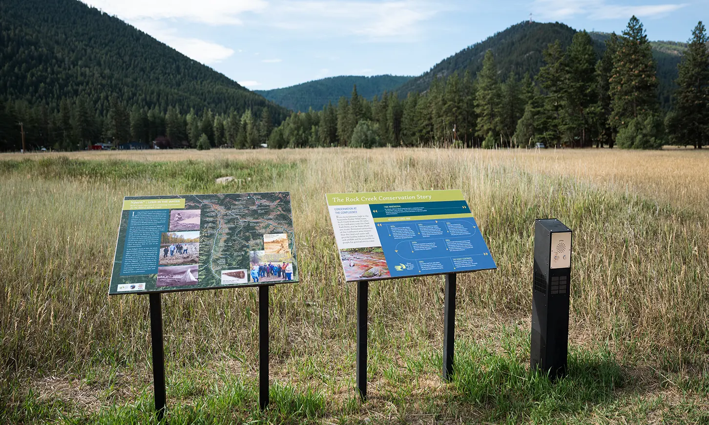 Two informational signs stand in a grassy meadow with mountains in the background.