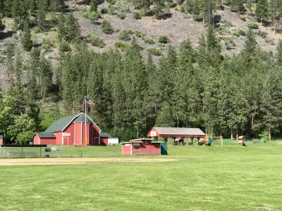 Photo from missoulacounty.gov A red barn-like building stands on a grassy field in front of a forested hillside.