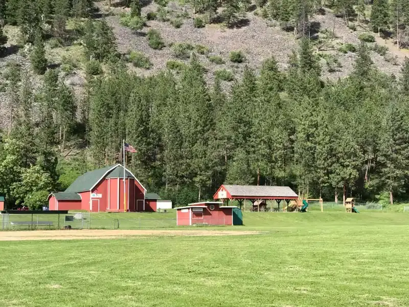 A red barn-like building stands on a grassy field in front of a forested hillside.