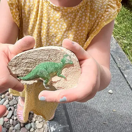 A child holds a piece of material featuring a small green toy dinosaur.
