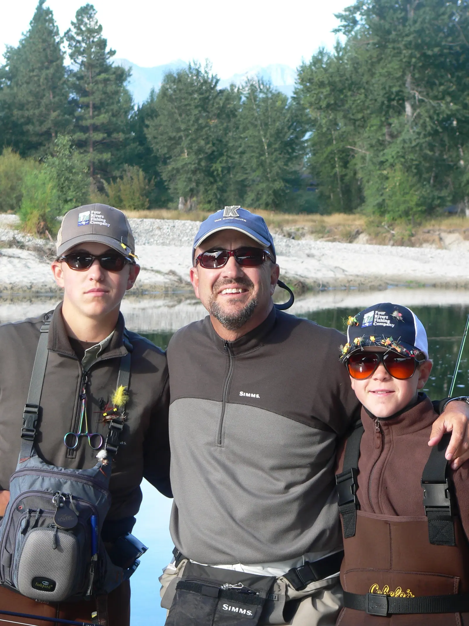 An adult man poses with two boys, all dressed in fishing gear, outdoors near a river.
