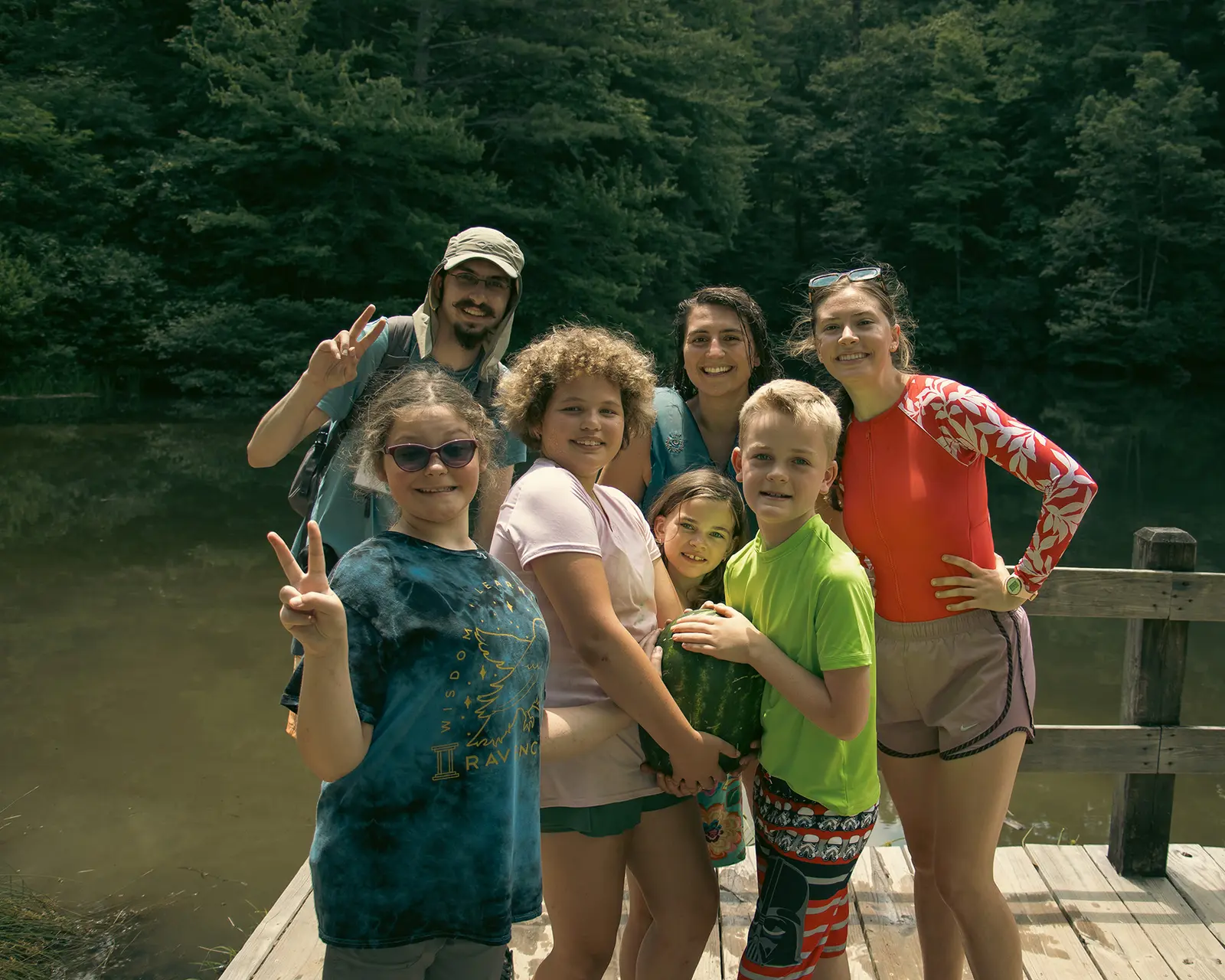 A group of children and adults pose for a picture on a wooden dock by a lake.