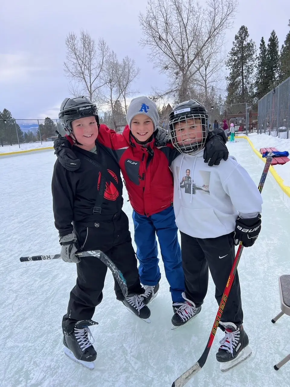 Three children stand on an ice rink wearing skates and hockey gear.