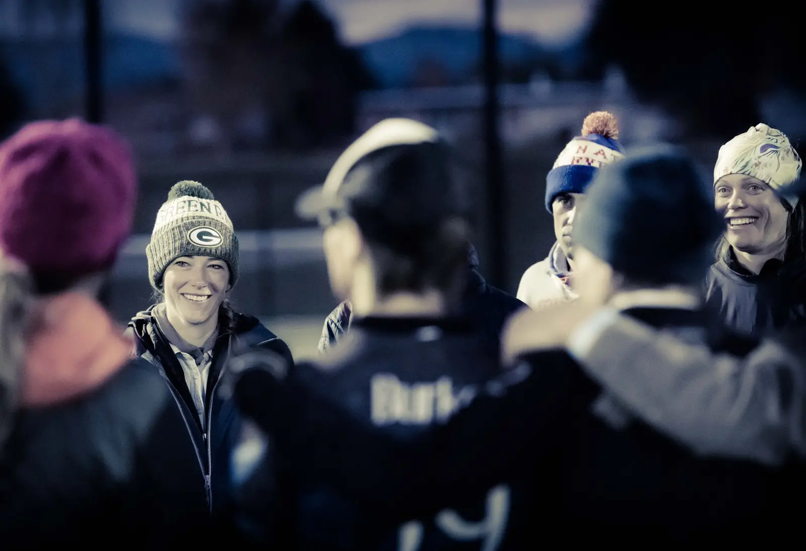 A group of people wearing winter hats stand together in a huddle outside.