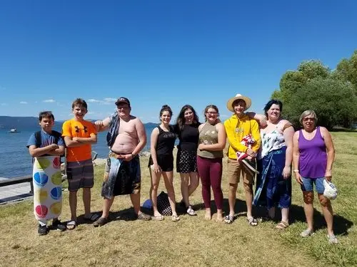 A group of young people and adults stand together outdoors near a lake on a sunny day.