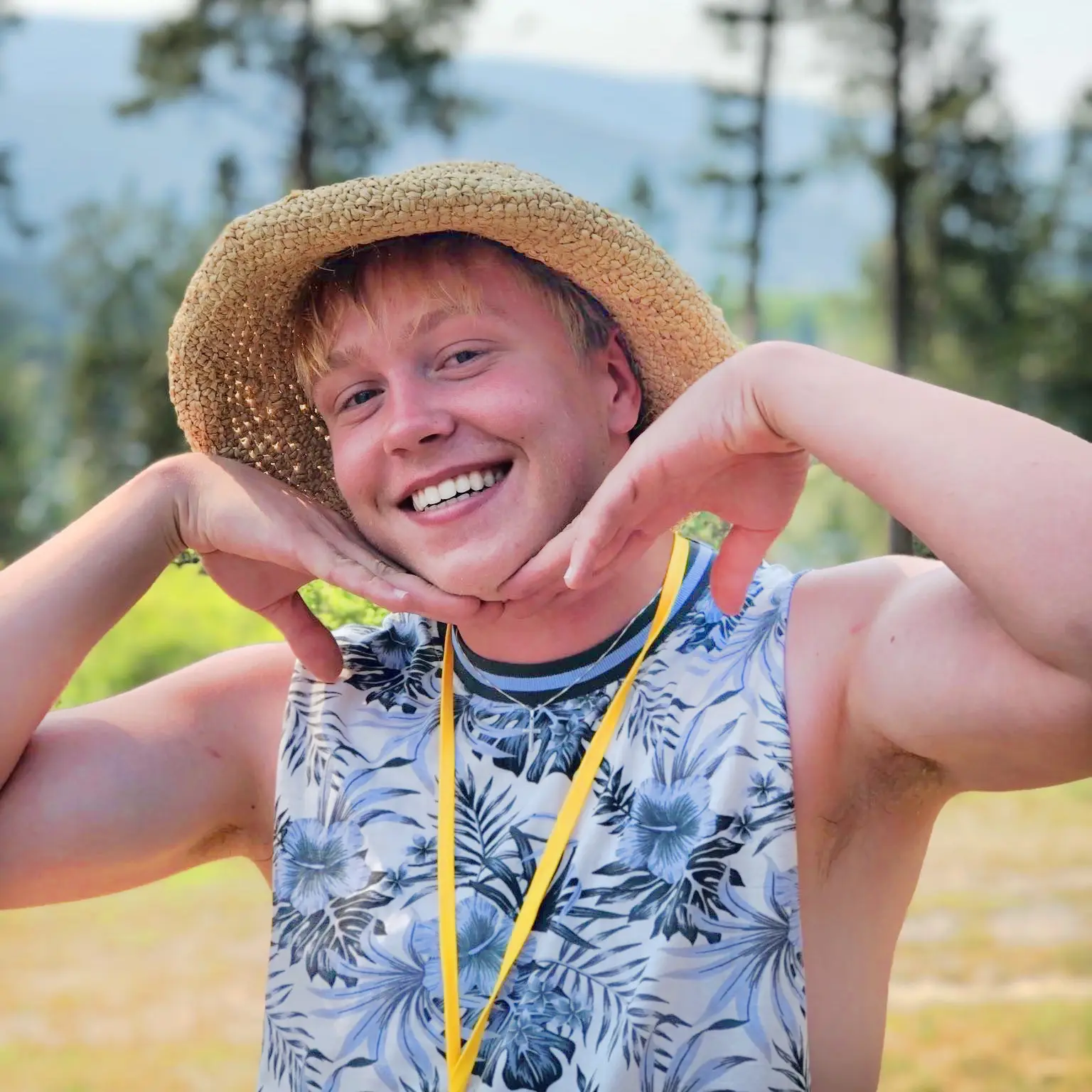 A smiling young adult wears a straw hat and floral print top outdoors.