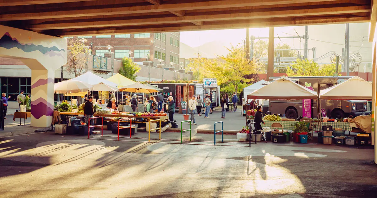 A sunny outdoor farmers market is set up under a bridge with various stalls and vendors.