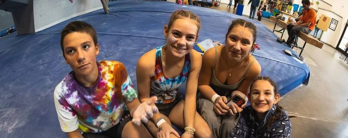 A group of young people with glitter on their faces sit together on a climbing gym mat.