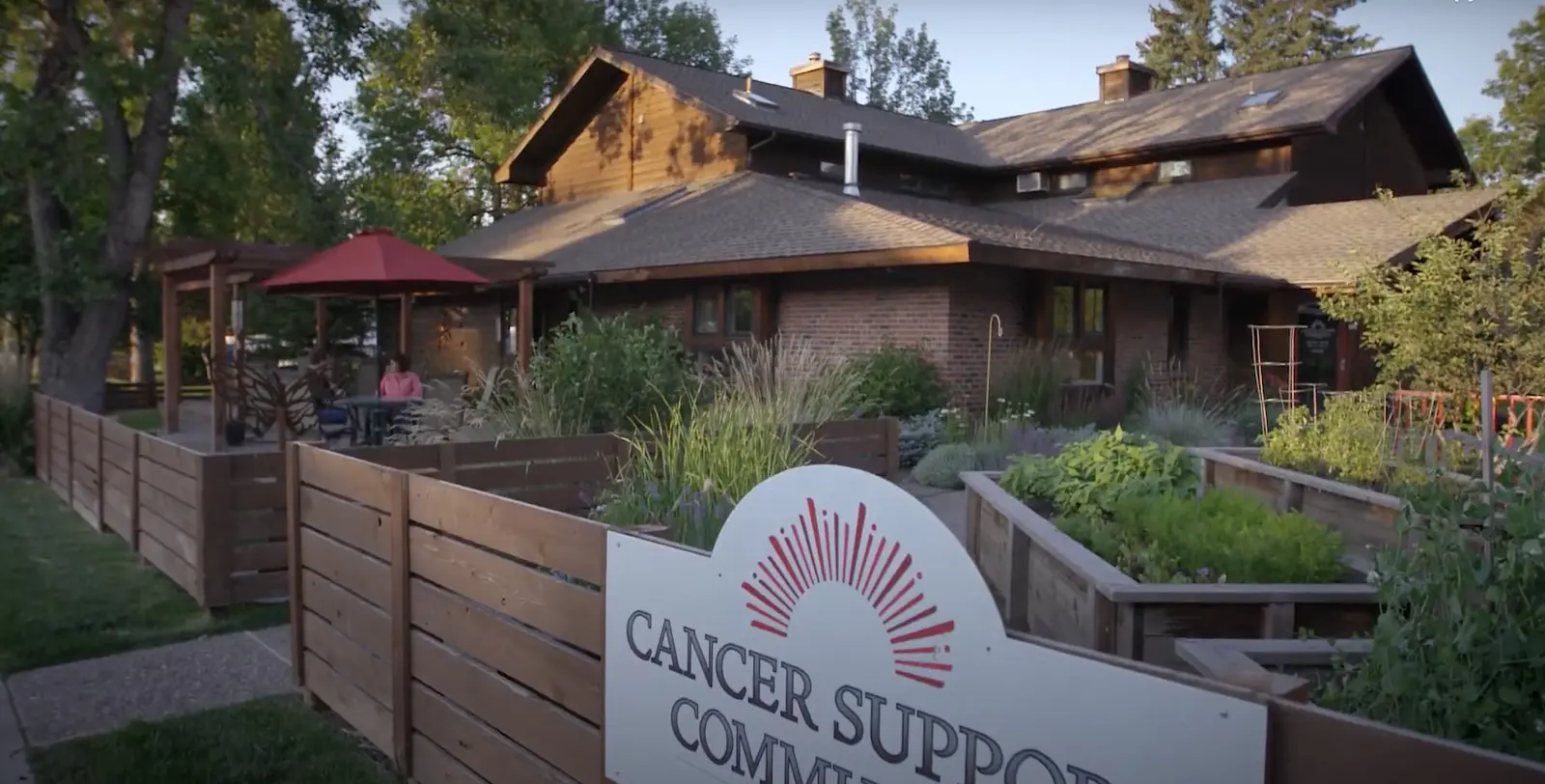 A wooden building surrounded by greenery and a garden features a sign for a cancer support community.