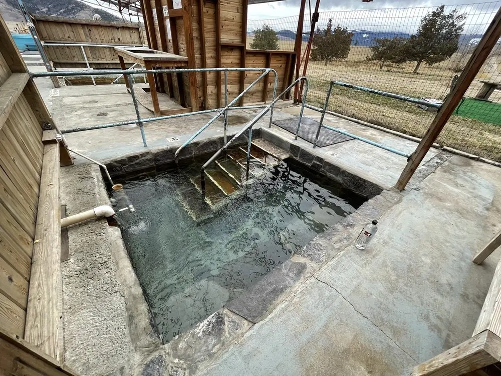 A stone-lined hot spring pool with metal handrails is situated outdoors under a wooden shelter.