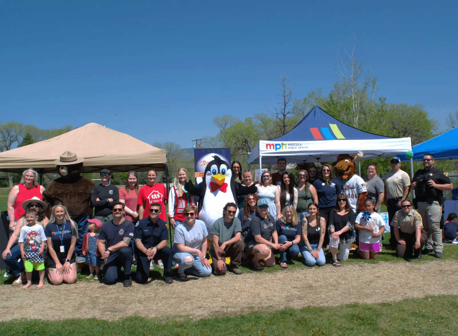 A large group of people and mascots pose for a photo in a park under a sunny sky.