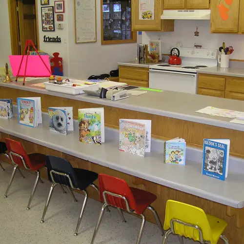 A row of children's books is displayed on a counter in a room with child-sized chairs.