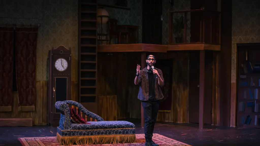 A man holding a microphone speaks while standing on a theatrical stage set.