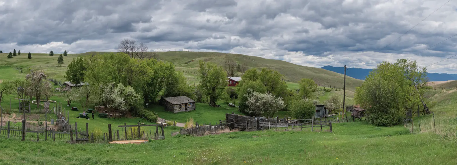 A wide-angle landscape view features historic wooden buildings nestled in a green valley under a cloudy sky.