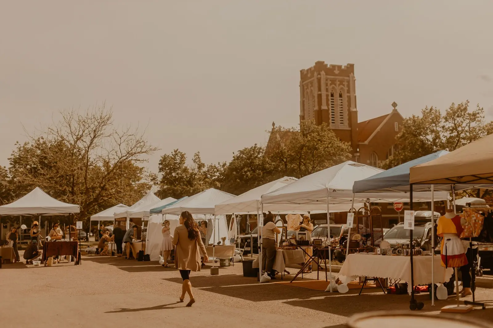 An outdoor market with white tents, vendors, and shoppers, set against a background of trees and a large brick church with a tall tower.