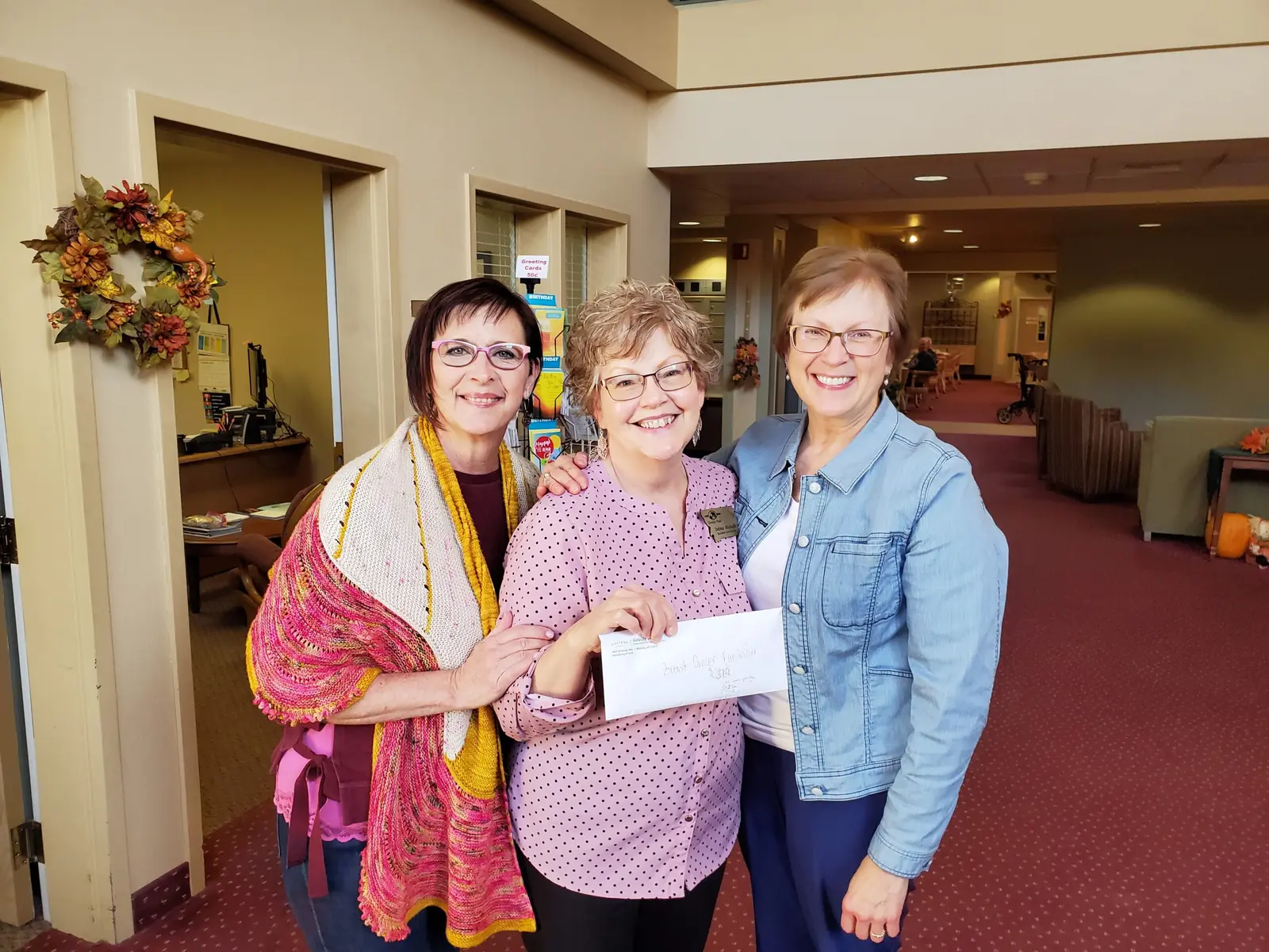 Three women pose together in an indoor setting while one holds an envelope.
