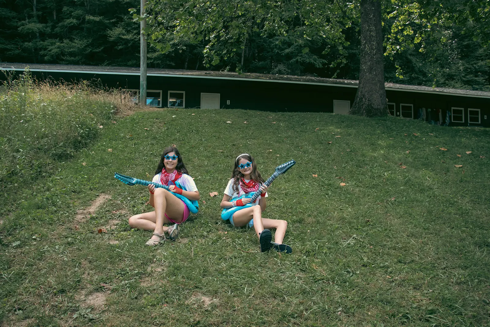 Two girls wearing star-shaped sunglasses sit on a grassy hillside holding inflatable guitars.
